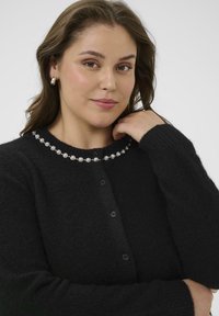 Gilet en tricot noir avec un col rond, orné de motifs floraux blancs. Il présente des boutons noirs et des poignets côtelés.