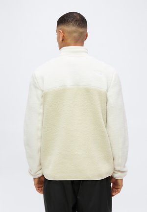 Hombre con chaqueta de forro polar bicolor beige y blanco roto visto desde atrás sobre un fondo claro y liso.