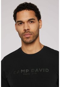 Camiseta negra de algodón suave, con un cuello redondo y un estampado de logo mate en tonos oscuros en el pecho.