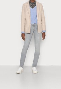 Blazer en laine beige sur un pull côtelé bleu clair, associé à un jean skinny gris clair et des baskets blanches. L'écharpe a un motif noir et blanc.