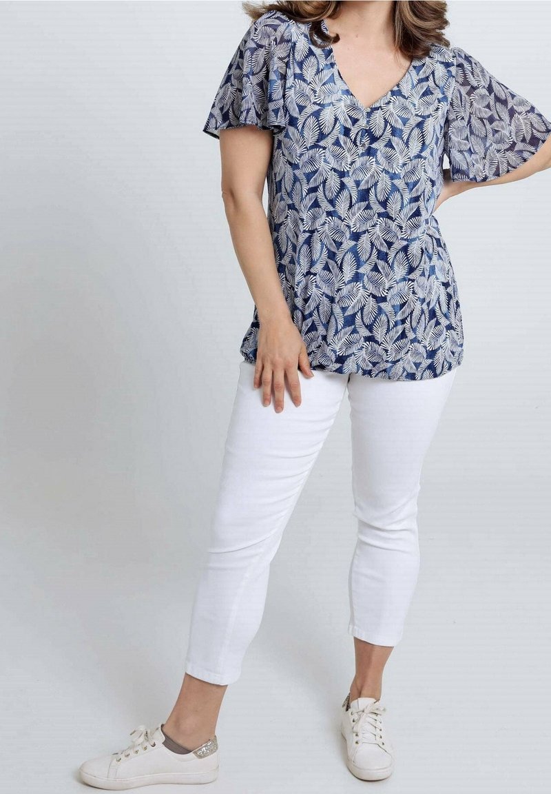 Marineblauwe blouse met wit bladmotief, V-hals en wijd uitlopende mouwen. Gecombineerd met witte slim-fit broek en witte sneakers.