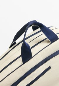 Vaaleabeige laukku, jossa on tummansinisiä raitakoristeita. Varustettu tukevilla tummansiniaisilla kangaskahvoilla ja useilla vetoketjullisilla taskuilla. Sileä pinta.