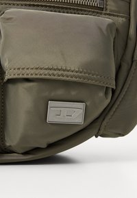 Poche de sac à dos en nylon vert avec un plaque logo argentée, des coutures visibles et une finition texturée, avec un détail de zip en haut.