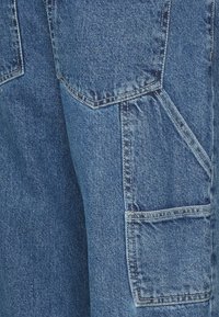Calças cargo de ganga azul com múltiplos bolsos, design de perna reta e detalhes de costura visíveis. O material tem um acabamento ligeiramente texturizado.
