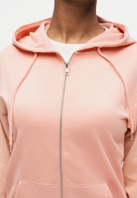 Άτομο που φοράει ροζ φούτερ με φερμουάρ πάνω από λευκό ριμπ μπλουζάκι, με το φερμουάρ του φούτερ μισάνοιχτο και την κουκούλα να ακουμπάει στους ώμους.