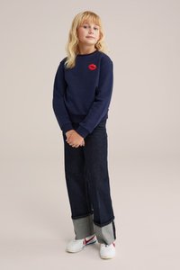 Mornarsko modra bluza s vezenjem rdečih ustnic, v kombinaciji s temnobluzimi širokimi kavbojkami z svetlo sivo obrobo. Bele superge dopolnjujejo oblačilo.