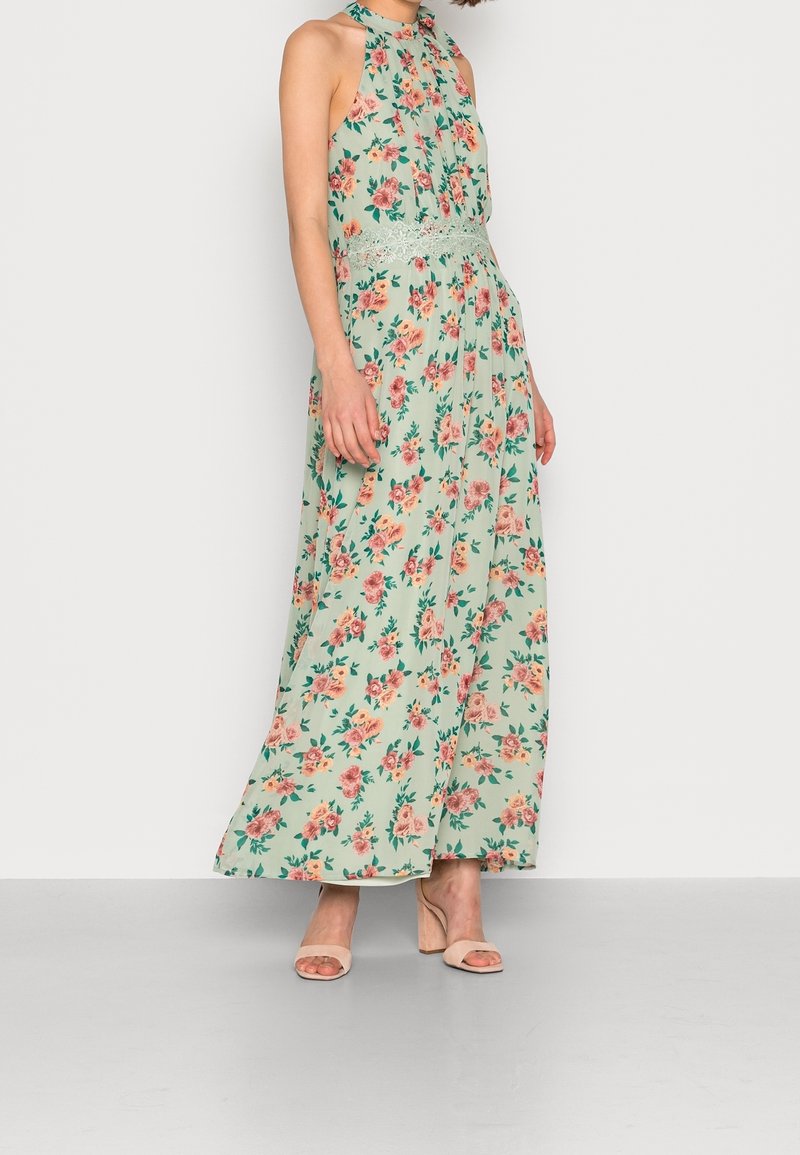 Femme portant une robe maxi sans manches vert menthe à imprimé floral rose et taille en dentelle, associée à des sandales à talons carrés nude.