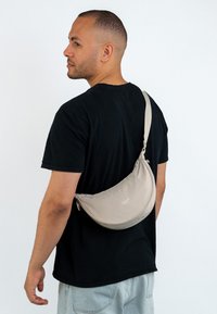 Uomo che indossa una maglietta nera e jeans in denim chiari, con una borsa a tracolla beige slacciata sulla schiena, davanti a uno sfondo semplice.