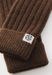 Brown ribbed knit gloves with a white tag featuring a black circular logo, laid on a white surface.