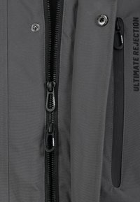 Gray jacket featuring multiple zippered pockets, smooth texture, and a bold "ULTIMATE REJECTION" print. Black hardware accents detail design.