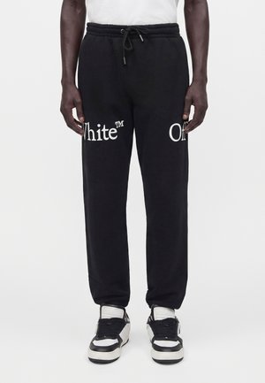 Homme portant un pantalon de survêtement noir avec le texte blanc "White™" et "Off" sur les cuisses, associé à des baskets noires et blanches, debout devant un fond blanc.