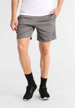 Mand iført grå atletikshorts, sorte sokker og sorte løbesko, stående med den ene hånd i lommen mod en hvid baggrund.