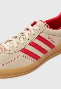 Baskets en daim beige avec trois rayures rouges et la marque "Gazelle", dotées d'une semelle en caoutchouc et de lacets blancs. Intérieur de couleur unie.