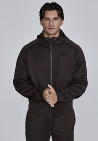 Donkerbruine zip-up hoodie met een capuchon, voorzien van een voork pocket en logo-detail op de borst. Gemaakt van glad, elastisch materiaal.