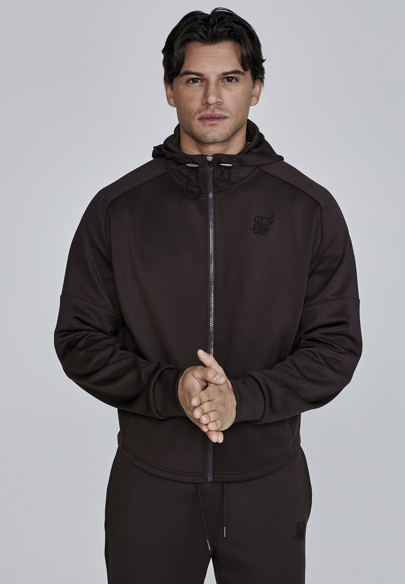 Donkerbruine zip-up hoodie met een capuchon, voorzien van een voork pocket en logo-detail op de borst. Gemaakt van glad, elastisch materiaal.