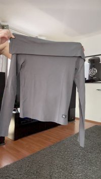 Mani che tengono una maglietta grigia a maniche lunghe e scollatura a spalle scoperte, con un piccolo logo Calvin Klein vicino all'orlo inferiore, in un soggiorno.