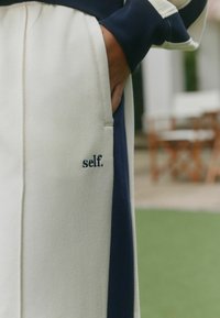 Šviesios spalvos, tekstūruotos sportinės kelnės su tamsiai mėlynomis šoninėmis juostelėmis, kišenėje išsiuvinėtu „self.“ logotipu. Laidnus kirpimas, su šoninėmis kišenėmis.