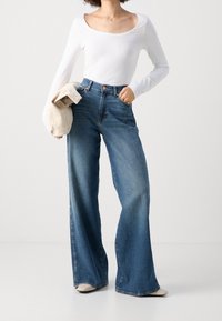 Vit långärmad topp som matchas med högmidjade, vida blåjeans som visar en klassisk denimfinish och lätt blekning.