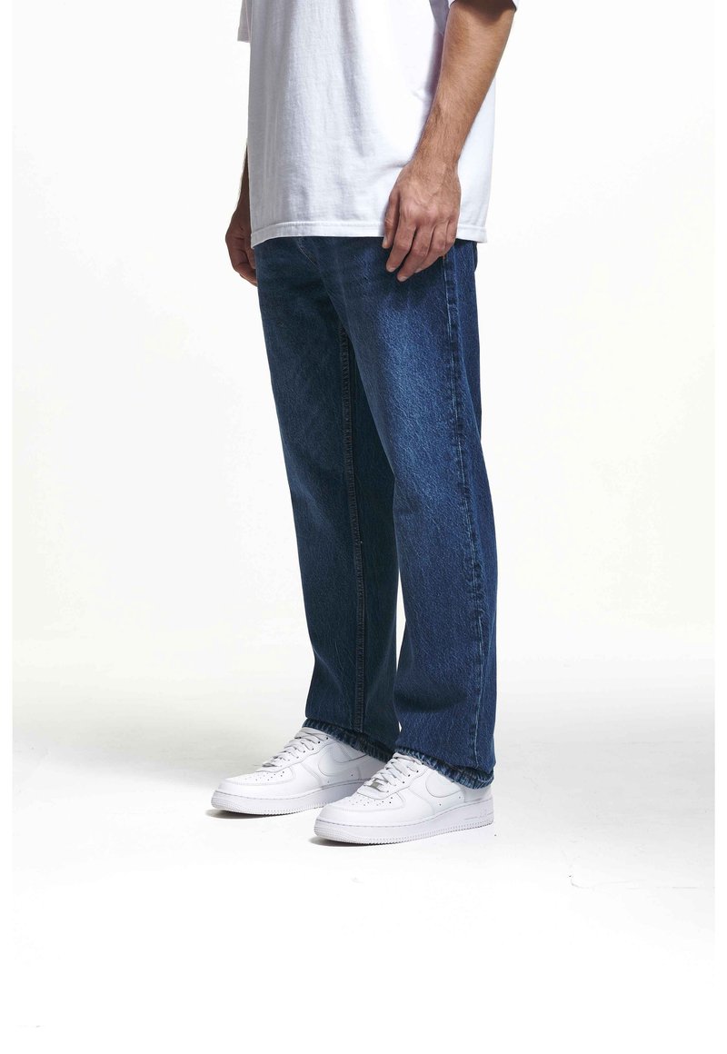 Niebieskie dżinsy z denimu o luźnym kroju, z podwiniętymi nogawkami i przednimi kieszeniami. Połączone z białymi trampkami, noszone z luźną białą koszulką.