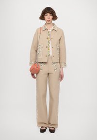 Beige embroidered jacket with pockets, paired with matching embroidered trousers. Textured fabric, floral accents, and a small orange bag.