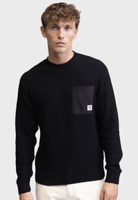 Pull en maille noir avec motif texturé, col rond et manches longues. Comprend une poche en tissu sombre avec détail de logo.