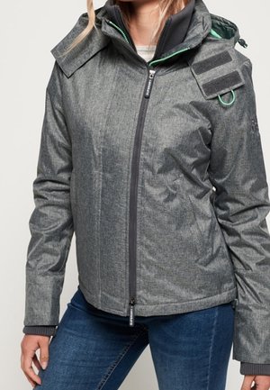 Kobieta ubrana w szarą kurtkę zapinaną na zamek z kapturem, rzepy na ramionach oraz niebieskie dżinsy, stoi z jedną ręką swobodnie opuszczoną wzdłuż ciała.