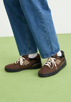 Person iført brune og sorte sneakers med hvide snørebånd og hvide sokker, kombineret med blå jeans, stående på en lysegrøn gulv.