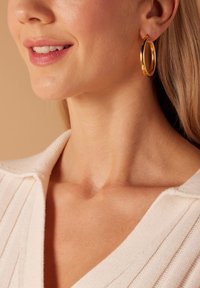 Boucle d'oreille en or présentant une surface lisse et polie. Le modèle porte un pull de couleur claire avec une texture côtelée et un décolleté en V.