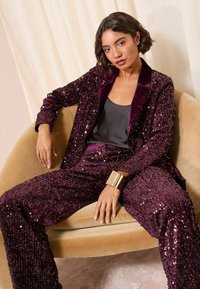 Set de sacou și pantaloni din paiete mov, cu un top din satin negru. Include o brățară masivă din aur. Purtați în timp ce erați așezat pe un scaun curbat.