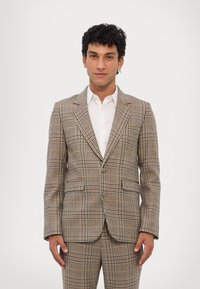 Barna és bézs kockás zakó, jellegzetes hajtókával, két elülső zsebbel és testreszabott szabással. Fehér ing felett viselve.