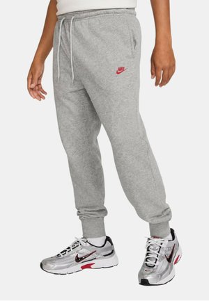 Parte inferiore del corpo di una persona che indossa pantaloni della tuta Nike grigio chiaro e sneaker Nike argento e nere con accenti rossi su sfondo bianco.