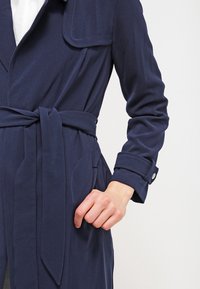 Gabardina azul marino con cinturón, cuello y bolsillos laterales. Presenta una textura suave y botones en los puños.