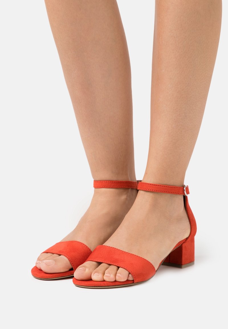 Pieds portant des sandales rouges à talons blocs ouvertes à l'avant avec des brides à la cheville sur fond blanc uni.