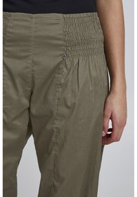 Pantaloni verde oliva chiaro con vita arricciata, design plissettato e un elegante accento argentato vicino alla vita. Tessuto morbido e leggero.