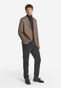 Brun kavaj över en svart polotröja, kombinerad med mörkgrå jeans och svarta skor. Kavajen har en knapp och ett strukturerat tyg.