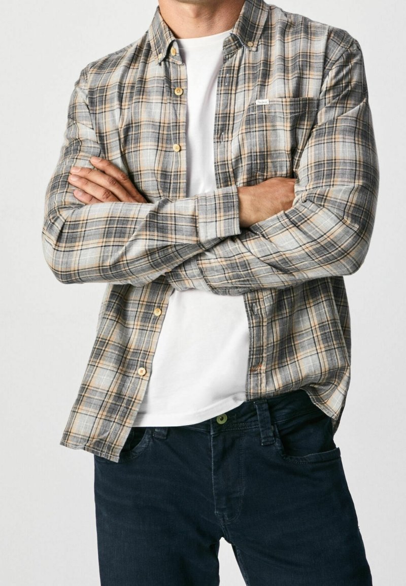 Camicia flanella a quadri nei toni grigi, blu e arancioni. Presenta un colletto con bottoni, una tasca sul petto ed è indossata sopra una maglietta bianca.
