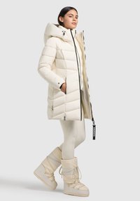 Svetlo krémová pufovaná bunda s kapucňou, s predným zipsom, diagonálnym prešívaním a čiernymi zipsovými detailmi. Vydaná s ladiacimi čižmami.