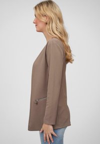 Haut long à manches avec texture marron, coupe décontractée et poche zippée sur le côté gauche, associé à un jean bleu clair.