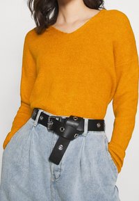 Senapsgul ribbad tröja med v-ringning, tillsammans med högmidjade ljusblå denimbyxor och ett svart läderbälte med detaljer i hårdvara.