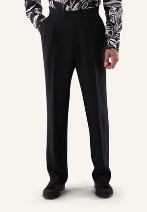 Mann trägt schwarze plissierte Hose, ein schwarz-weiß geblümtes Langarmhemd und dunkelbraune Wildleder-Loafer mit Quasten.