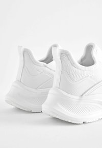 Weiße Sportschuhe mit einem Obermaterial aus Mesh, geschwungenem Fersendesign, strukturierten Akzenten und einer dicken Sohle mit Profilmuster.
