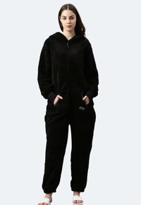 Schwarzer Fleece-Overall mit Kapuze, frontalen Taschen und elastischen Bündchen. Strukturierter Stoff, Reißverschluss und ein Logo-Detail auf der Tasche.