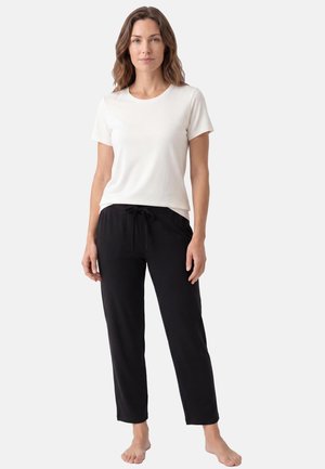 Femme debout pieds nus portant un t-shirt blanc uni à manches courtes et un pantalon noir à cordon sur un fond clair.