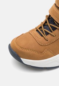 Zapatillas de deporte de cuero sintético marrón con lengua de malla azul marino, cordones en contraste y entresuela blanca. Presenta costuras visibles y una superficie texturizada.