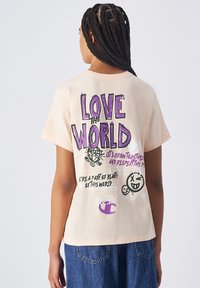 Lichtbeige katoenen t-shirt met korte mouwen. Grafisch ontwerp op de achterkant met paarse en zwarte tekst, een glimlachend gezicht en een illustratie van een wereldbol.