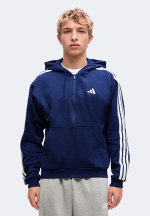 Jeune homme aux cheveux blonds courts portant un sweat à capuche zippé bleu marine avec des bandes blanches sur les manches et un pantalon de jogging gris.