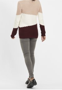 Pull en maille épaisse avec des blocs de couleurs horizontaux en beige, crème et bordeaux. Associé à un pantalon gris ajusté et des chaussures noires.