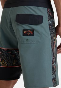 Pánske plavky v odtieni teal s čiernym bočným panelom zdobeným zložitými vzormi paisley. Obsahujú čiernu nášivku na vrecku a logo.