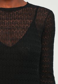 Haut noir en tricot présentant un motif texturé, une coupe ajustée et des manches longues avec un décolleté rond. Le matériau semble doux et léger.