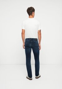 Jeans de mezclilla azul oscuro con un ajuste slim, que cuentan con un diseño estándar de cinco bolsillos y un parche de logo en la parte trasera de la cintura. Llevados con una camiseta blanca.